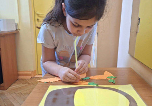 Zdjęcie przedstawia Olę z grupy IV wykonującą ćwiczenie oddechowe - przenoszenie papierowych marchewek do koszyka za pomoca słomki do napojów.