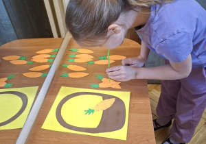 Zdjęcie przedstawia Klarę J. z grupy IV wykonującą ćwiczenie oddechowe - przenoszenie papierowych marchewek do koszyka za pomoca słomki do napojów.