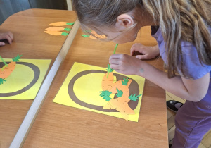 Zdjęcie przedstawia Klarę J. z grupy IV wykonującą ćwiczenie oddechowe - przenoszenie papierowych marchewek do koszyka za pomoca słomki do napojów.