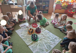 Na tym zdjęciu widać siedzące w kole dzieci oraz Emmę, która umieszcza nasionko fasoli w kubeczku wypełnionym ziemią.
