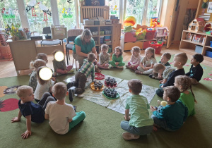 Na tym zdjęciu widać siedzące w kole dzieci oraz Adasia, który umieszcza nasionko fasoli w kubeczku wypełnionym ziemią.