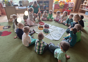 Na tym zdjęciu widać siedzące w kole dzieci oraz Lenę T., która umieszcza nasionko fasoli w kubeczku wypełnionym ziemią.