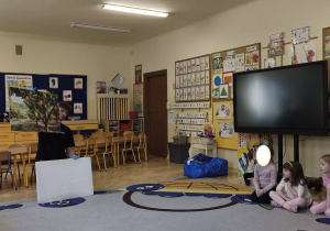 Zdjęcie przedstawia prowadzącego warsztaty, który opowiada dzieciom, w jaki sposób powstaje czekolada.