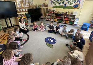 Zdjęcie przedstawia wszystkie dzieci siedzące w kole na dywanie oraz prowadzącą warsztaty.