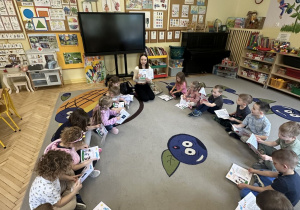 Zdjęcie przedstawia dzieci siedzace w kole podczas oglądania i omawiania kart pracy oraz prowadzącą zajęcia.