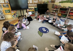 Zdjęcie przedstawia dzieci siedzace w kole podczas oglądania i omawiania kart pracy oraz prowadzącą zajęcia.