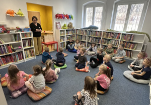 Zdjęcie przedstawia dzieci siedzace na poduchach i słuchające opowieści pani bibliotekarki.
