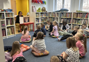Zdjęcie przedstawia dzieci siedzace na poduchach i słuchające bajki czytanej przez panią bibliotekarkę.