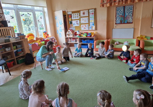 Zdjęcie przedstawia dzieci siedzące na dywanie w kole i mamę Damiana czytającą dzieciom pierwszą z książeczek.