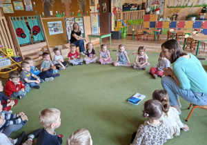 Zdjęcie przedstawia dzieci siedzące na dywanie w kole i mamę Damiana czytającą dzieciom pierwszą z książeczek.