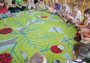 Zdjęcie przedstawia dzieci siedzące w kole na dywanie oraz prowadzącą warsztaty.