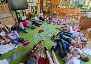 Zdjęcie przedstawia dzieci leżace w kole na dywanie oraz prowadzącą warsztaty.