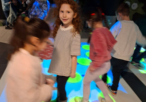 Zdjęcie przedstawia Mayę z grupy IV z koleżankami bawiące się na dywanie interaktywnym.