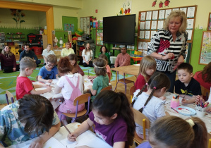 Zdjęcie przedstawia dzieci siedzące przy stolikach podczas wykonywania pracy plastycznej oraz obserwujących ich rodziców.