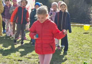 Zdjęcie przedstawia jedną z dziewczynek przenoszącą jajko na łyżce oraz pozostałe dzieci stojące z tyłu.