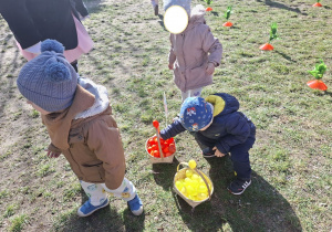 Zdjęcie przedstawia troje dzieci przynoszące jajka do koszyczka.