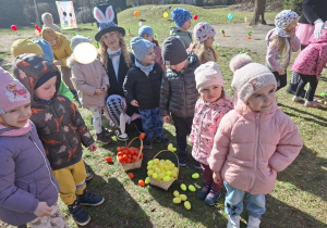 Zdjęcie przedstawia dzieci stojące obok koszyczków z zebranymi w ogrodzie jajkami.