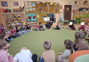 Zdjęcie przedstawia mamę Mikołaja czytającą książeczkę oraz dzieci siedzące w kole, słuchające opowieści.