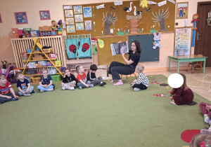 Zdjęcie przedstawia mamę Mikołaja czytającą książeczkę oraz dzieci siedzące w kole, słuchające opowieści.