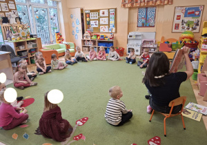 Zdjęcie przedstawia mamę Mikołaja czytającą książeczkę oraz dzieci siedzące w kole, słuchające opowieści.