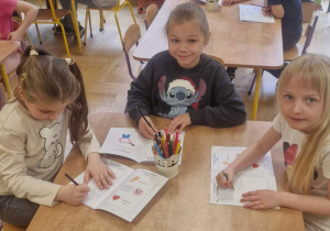 Zdjęcie przedstawia dzieci siedzące przy stolikach, wykonujące zadanie w kartach pracy.