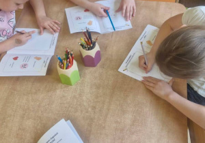 Zdjęcie przedstawia dzieci siedzące przy stolikach, wykonujące zadanie w kartach pracy.