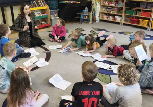 Na tym zdjęciu widać prowadzącą zajęcia oraz dzieci siedzące w kole podczas omawiania kart pracy.