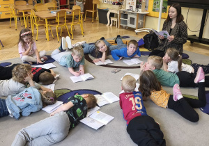 Zdjęcie przedstawia dzieci leżące na dywanie oraz prowadzącą, która czyta dzieciom opowiadanie.