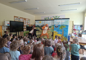 Zdjęcie przedstawia troje dzieci wraz z jednym z bohaterów na scenie oraz przedszkolaki oglądajace bajkę.