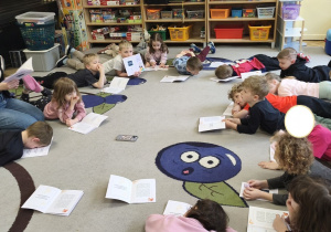 Zdjęcie przedstawia leżące na dywanie oraz prowadzącą warsztaty, która czyta dzieciom opowidanie o smutku.