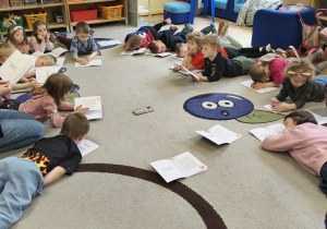 Zdjęcie przedstawia leżące na dywanie oraz prowadzącą warsztaty, która czyta dzieciom opowidanie o smutku.