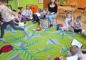 Na tym zdjęciu widać dzieci leżące na dywanie w kole, słuchające opowiadania czytanego przez prowadzącą.