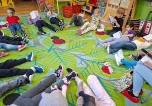 Na tym zdjęciu widać dzieci leżące na dywanie w kole, słuchające opowiadania czytanego przez prowadzącą.