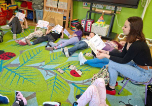 Na tym zdjęciu widać dzieci leżące na dywanie w kole, słuchające opowiadania czytanego przez prowadzącą.