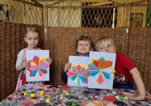 Zdjęcie przedstawia troje dzieci prezentująch prace - kwiatki wykonane metodą origami z kwadratu.