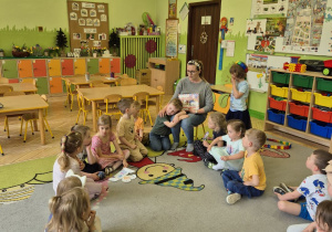 Zdjęcie przedstawia mamę Adasia czytającą dzieciom bajkę oraz dzieci siedzące na dywanie.