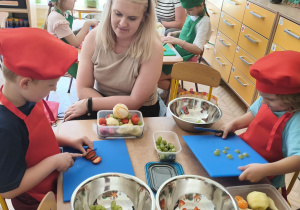 Zdjęcie przedstawia Mikołaja oraz Adasia W. z grupy III z mamą krojących owoce w drobną kostkę.