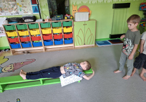 Zdjęcie przedstawia Filipa L. z grupy IV wykonującego ćwiczenia mięśni grzbietu oraz obręczy barkowej z wykorzystaniem ławeczki gimnastycznej oraz Tymona z grupy III oczekującego na swoją kolej.