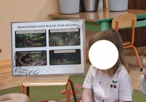 Zdjęcie przedstawia jedną z dziewczynek siedzącą obok planszy ukazującej naturalne środowisko zwierzątka.