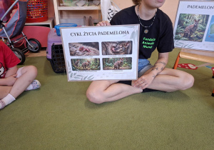 Na tym zdjęciu widać prowadzącą, która w parciu o planszę opowiada o cyklu życia zwierzątka.