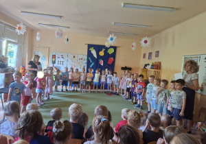 ZDjęcie przedstawia dzieci z grupy I i II stojące w półkolu podczas występu.