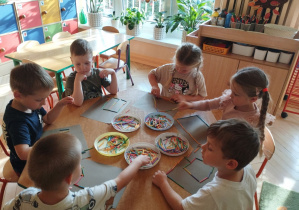 Zdjęcie przedstawia Antosia, Adasia, Bartka, Henia P., Kamilkę oraz Lenkę T. z grupy II siedzących przy stoliku, którzy układają wzory z małych, kolorowych patyczków.