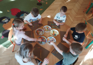 Zdjęcie przedstawia Antosia, Adasia, Bartka, Henia P., Kamilkę oraz Lenkę T. z grupy II siedzących przy stoliku, którzy układają wzory z małych, kolorowych patyczków.