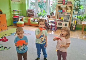 Zdjęcie przedstawia troje dzieci z grupy I wykonujących ćwiczenia oddechowe z wykorzystaniem papierowych liści.