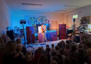 Zdjęcie przedstawia jedną ze scen teatrzyku oraz dzieci siedzące na dywanie i oglądające przedstawienie.
