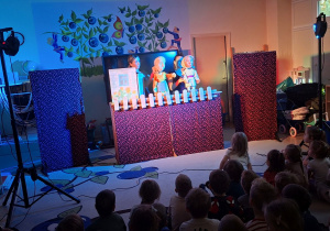 Zdjęcie przedstawia jedną ze scen teatrzyku oraz dzieci siedzące na dywanie i oglądające przedstawienie.