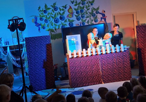 Zdjęcie przedstawia jedną ze scen teatrzyku oraz dzieci siedzące na dywanie i oglądające przedstawienie.