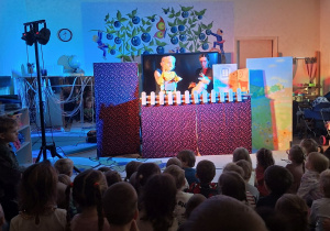 Zdjęcie przedstawia jedną ze scen teatrzyku oraz dzieci siedzące na dywanie i oglądające przedstawienie.