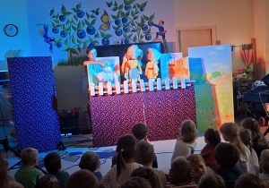 Zdjęcie przedstawia jedną ze scen teatrzyku oraz dzieci siedzące na dywanie i oglądające przedstawienie.